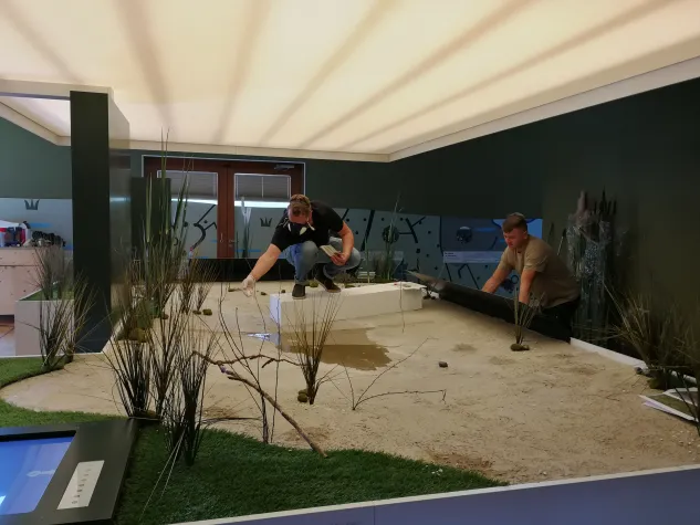2 Mitarbeiter gestalten Bereich eines Besucherzentrums, Fläche mit Sand, Pflanzen und Wasser