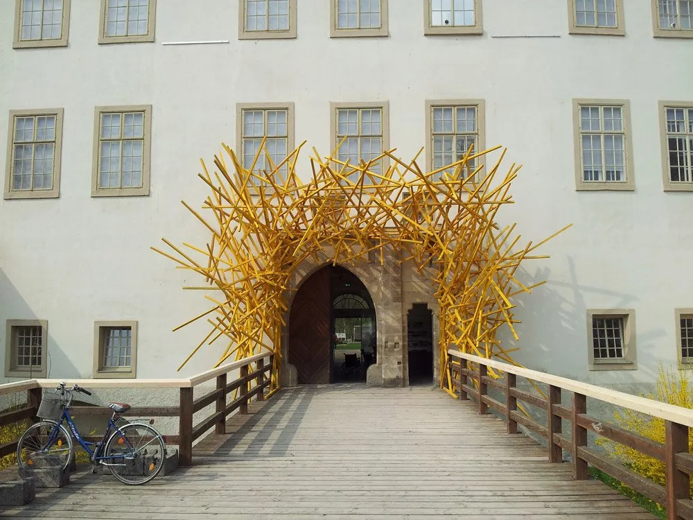 Ausstellungsraum: Holzbrücke mit gelber Skulptur über dem Eingang eines weißen Gebäudes, ein Fahrrad steht seitlich, Fenster mit grauen Rahmen im Hintergrund.