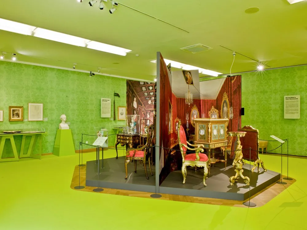 Ausstellungsraum: Grüne Wände und Boden, Vitrinen mit antiken Möbeln und Statuen, Wandbilder mit historischen Szenen