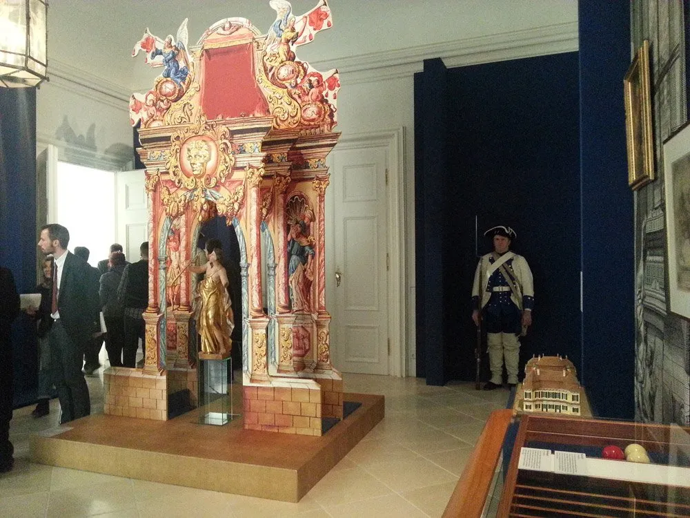 Ausstellungsraum: Prächtiger roter und goldener Altar mit Statue, umgeben von Besuchern, ein historisch gekleideter Wärter, in einem Raum mit blauen Wänden und Marmorboden