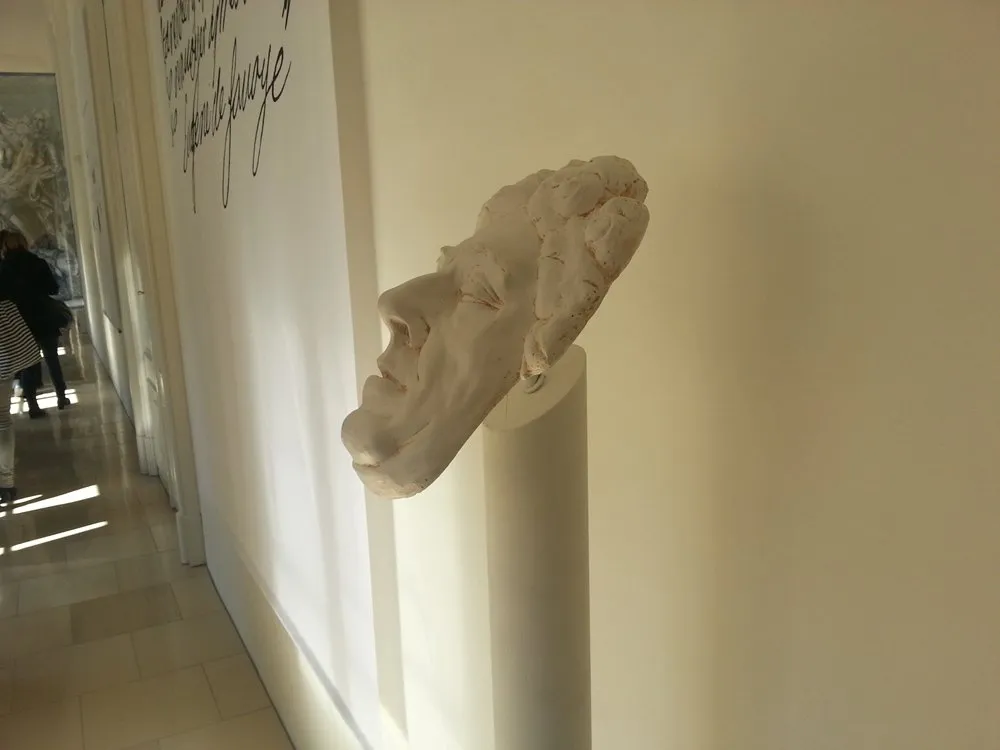 Gesichtsskulptur auf weißem Sockel, im Hintergrund Besucher und Text an der Wand