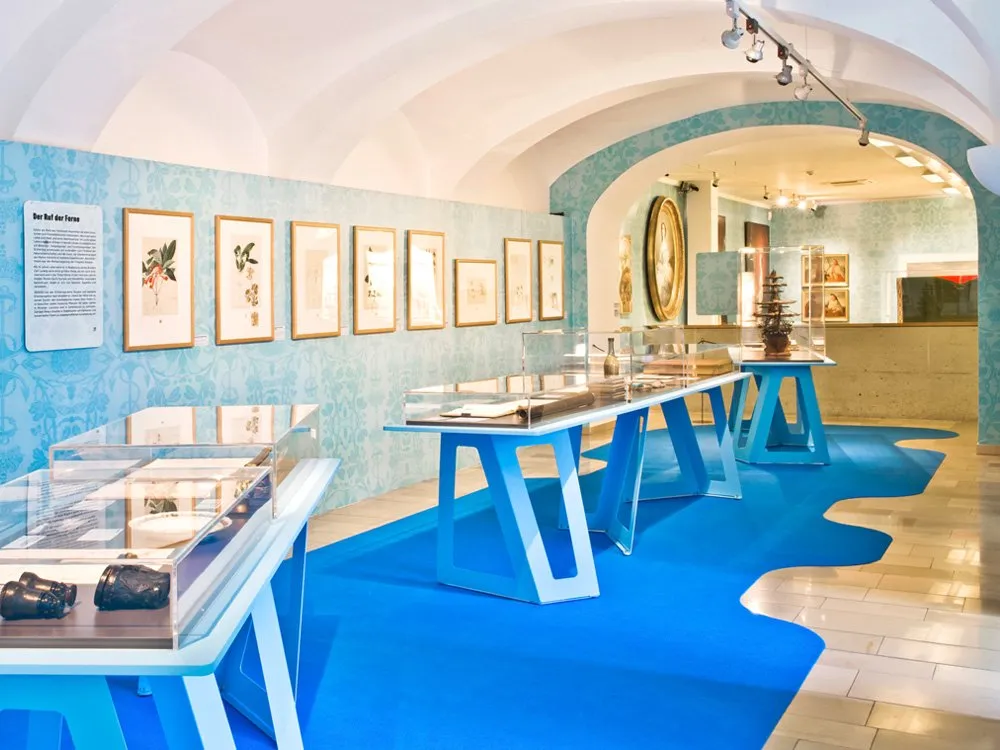 Ausstellungsraum: Blauer Raum mit gewölbter Decke, Vitrinen mit Artefakten und Bildern, großes goldgerahmtes Porträt an der Wand.