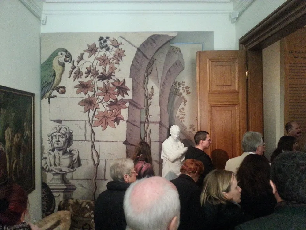 Ausstellungsraum: Wand mit floralem Muster, Papagei und Büste, eine weiße Statue im Zentrum, umgeben von Besuchern