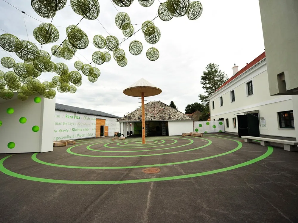 Ausstellungsraum: Offener Platz mit grünen Spirallinien, hängenden grünen Kugeln, einem Schirm mit steinernem Sockel und weißen Gebäuden im Hintergrund