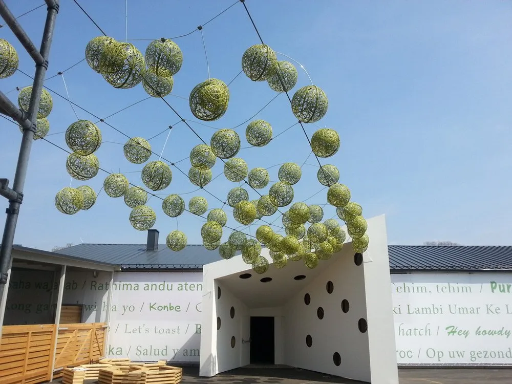 Ausstellungsraum: Offener Platz mit hängenden grünen Kugeln, weißem Gebäude mit grünen Texten und Punkten, Holzbauten und blauem Himmel im Hintergrund.