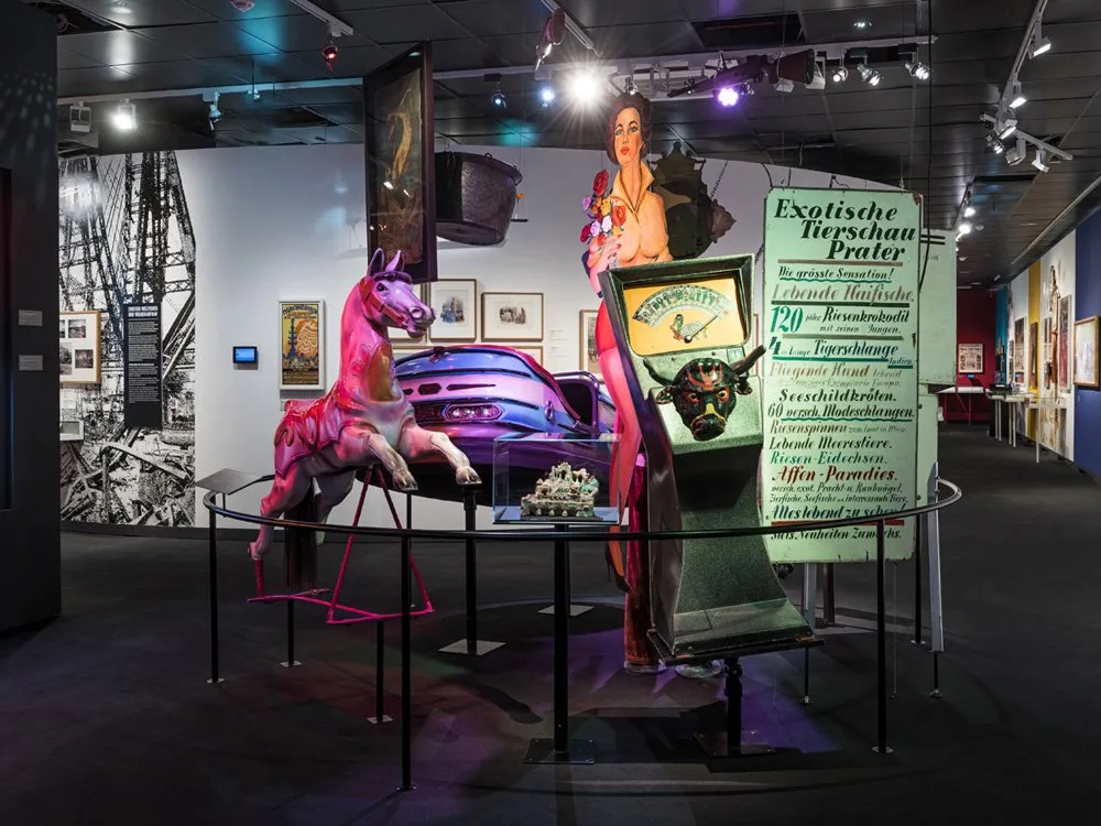 Ausstellungsraum: Pinkes Karussellpferd, blaues Auto-Modell, große Figur mit Blumen, Waage, Poster an der Wand
