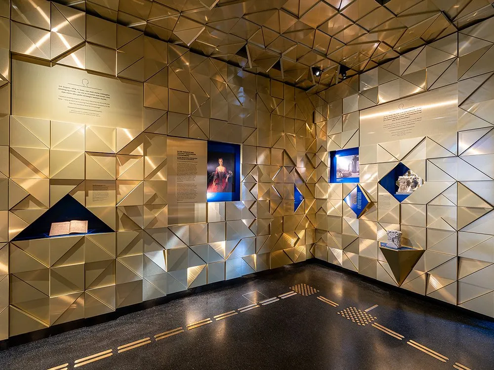 Ausstellungsraum mit goldenen geometrischen Wänden, Vitrinen mit historischen Exponaten und einem Porträt