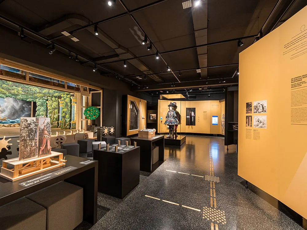 Ausstellungsraum mit interaktiven Stationen, einer Figur in historischem Gewand, Vitrinen mit Exponaten und Bildern an der Wand