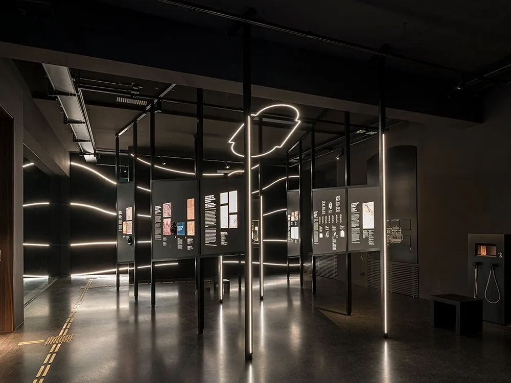 Ausstellungsraum mit schwarzen Panels, beleuchtete Displays mit Bildern und Beschriftungen, leuchtende Neonlinien an der Wand