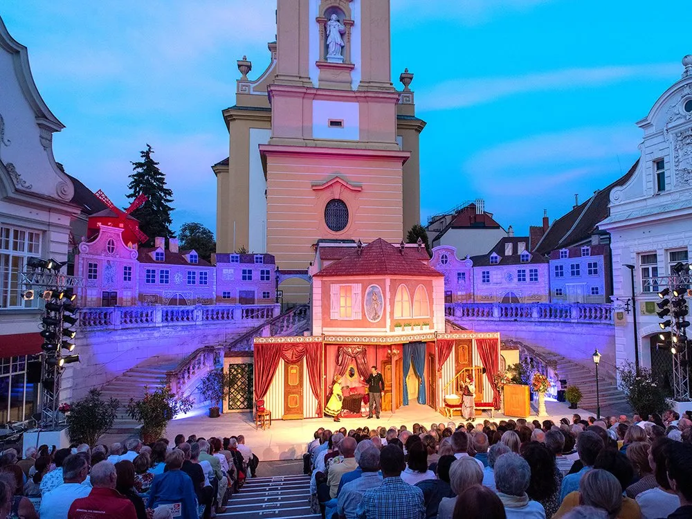 eiluftbühne: Theateraufführung mit Kulisse vor einer Kirche, Schauspieler auf der Bühne, Publikum auf Sitzreihen, beleuchtet bei Abenddämmerung