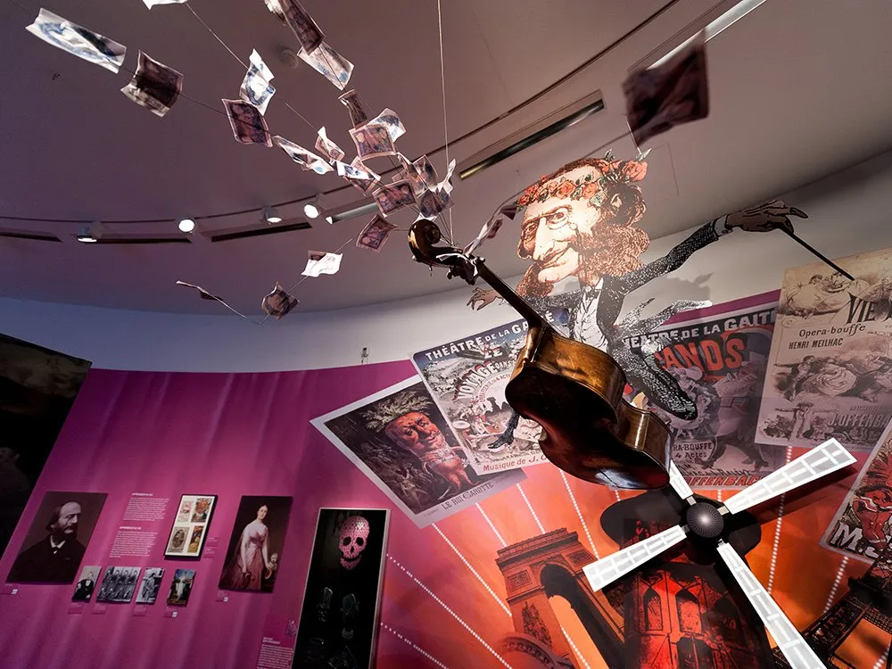 Ausstellungsraum: Pinker Raum mit hängenden Papierfetzen und einem Kontrabass, umgeben von Plakaten, mit Schädeldarstellungen und historischen Bildern an den Wänden