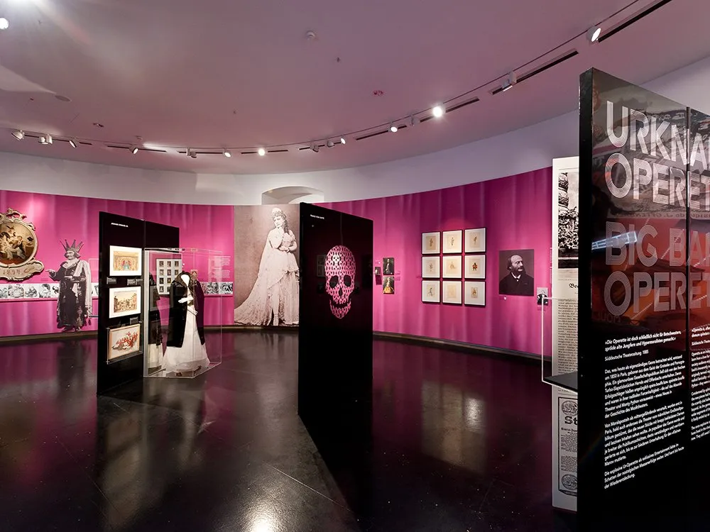 Ausstellungsraum: "Urknall Big Bang Operetta" mit historischen Kunstwerken, einem Kleid in einer Vitrine, einem rosa Schädel-Design und Fotos an einer pinken Wand