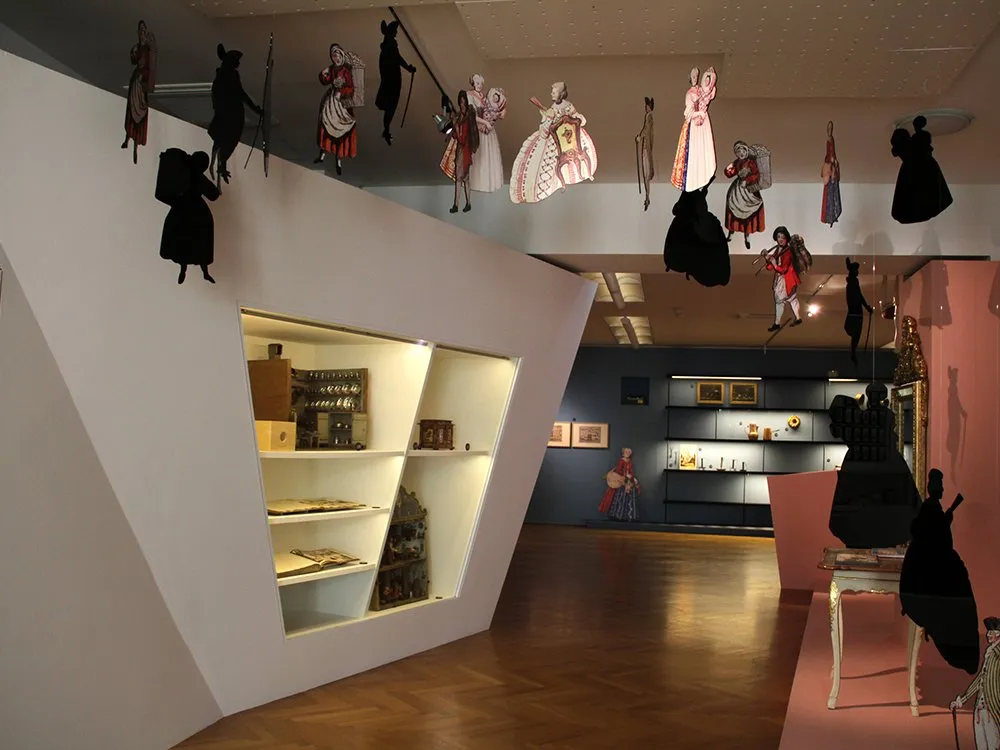 Ausstellungsraum: Weiße Vitrine mit historischen Gegenständen. Decke mit Pappfiguren und Schattenrisse