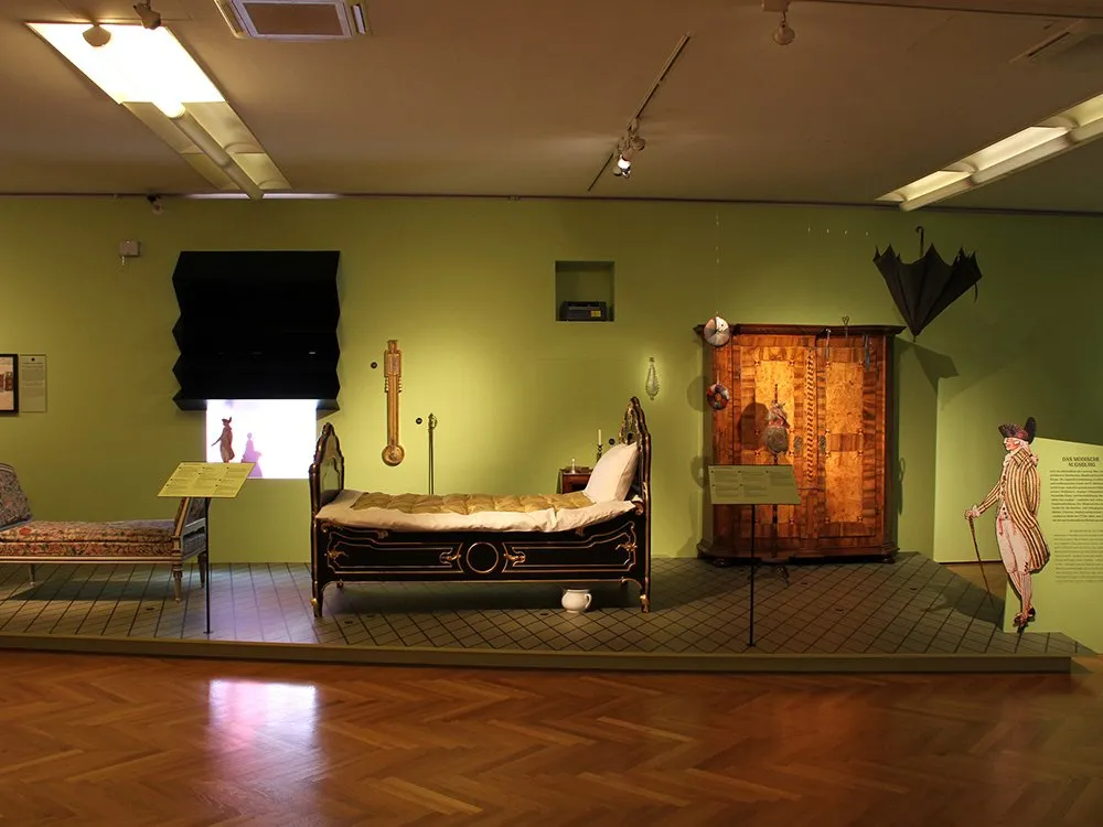 Ausstellungsraum: Grüne Wand mit historischem Bett, Schrank und Chaiselongue. Dekoriert mit Uhr, Schirm und einer Figur mit Stock