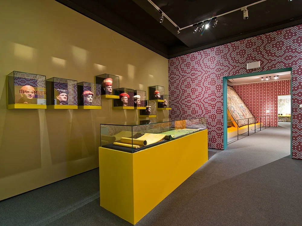 Ausstellungsraum mit gelben Wänden, einer Vitrine mit Dokumenten und mehreren bemalten Porträtköpfen in Vitrinen