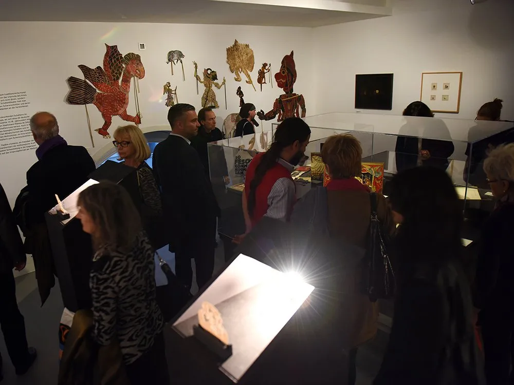 Ausstellung mit Besuchern, die traditionelle Schattenspielpuppen an der Wand und Ausstellungsstücke in Vitrinen betrachten