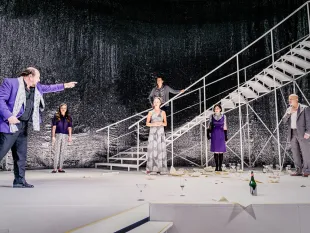Szene: Theaterbühne mit weißer Treppe und glitzerndem Hintergrund. Fünf Schauspieler, einer in lila Anzug zeigt dramatisch. Gläser, Champagnerflasche und Papiere liegen verstreut.