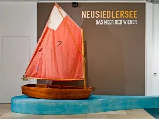 Modell eines Segelboots mit der Aufschrift "Neusiedlersee – Das Meer der Wiener" auf einer blauen Plattform