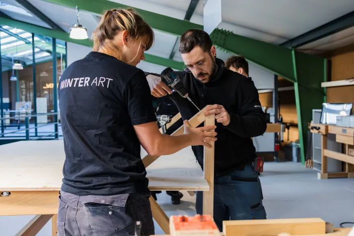 Werkstattraum mit zwei Mitarbeitern, die mit einer Bohrmaschine an einem Holzrahmen arbeiten