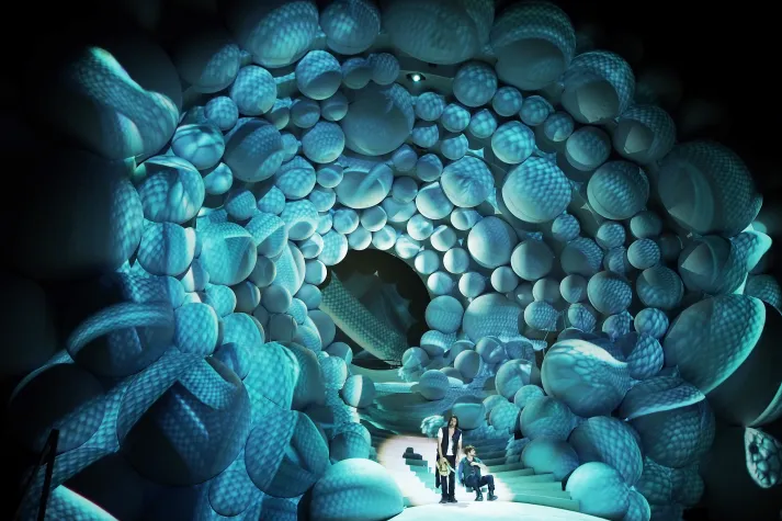 Bühnenbild: Drei Personen in einem Tunnel aus blauen, kugelförmigen Strukturen, die einen himmlischen Wolkenstrudel darstellen