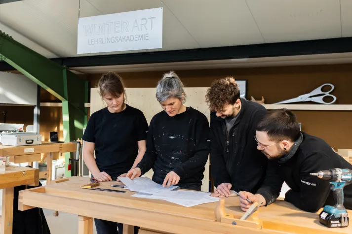 Lehrlingsausbildnerin steht zusammen mit 3 Lehrlingen hinter einer Hobelbank und schauen zusammen Pläne an