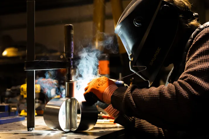 Nahaufnahme von Schlosser beim schweißen. Er trägt einen Helm und Handschuhe, Rauch steigt über dem Werkstück auf