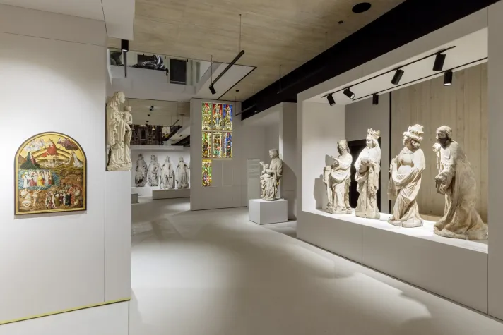 Ausstellungsraum mit Vitrinen, die historische Statuen und ein bunten Kirchenfenstern