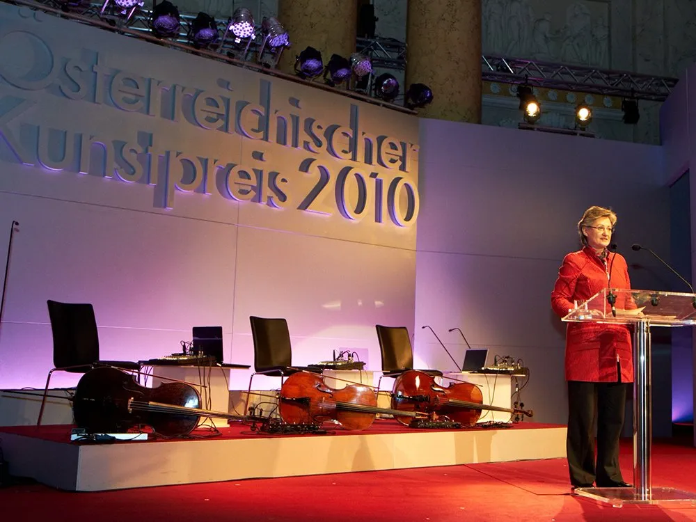 3D Schrift "Österreichischer Kunstpreis 2010" an Bühnenrückwand, Frau an Pult, Streichinstrumente neben Sessel am Boden