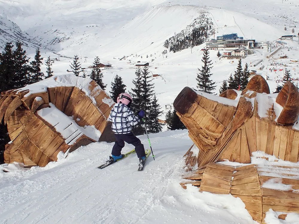 Zweiteilige Bärenskulptur, eine Seite: Kopf; andere Seite: Pfoten; schmale Piste dazwischen mit Kind auf Skia