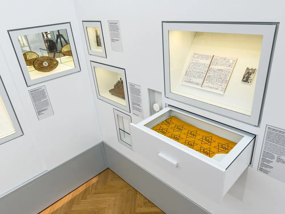 Ausstellungsraum: verschiedene Vitrinen mit historischen Artefakten, einem offenen Buch und einem gelben Tuch mit Davidsternen enthält