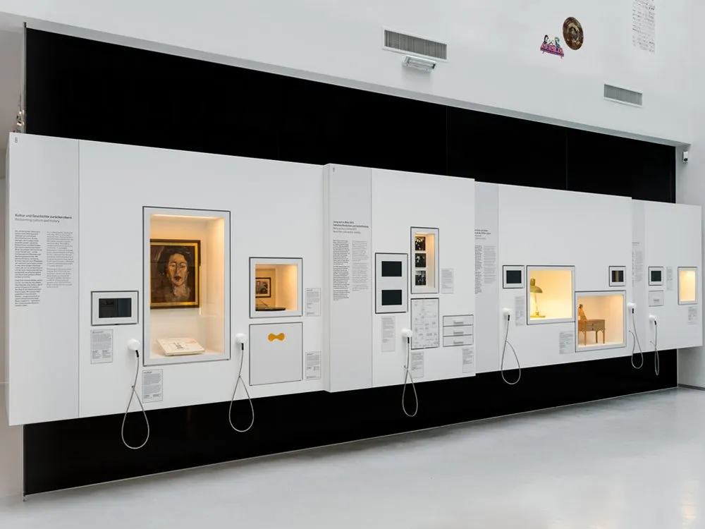 Ausstellungsraum mit weißen Wänden, die Vitrinen mit einem Gemälde, Büchern, einem Stuhl, einer Lampe und anderen Artefakten enthalten, sowie Audioguides