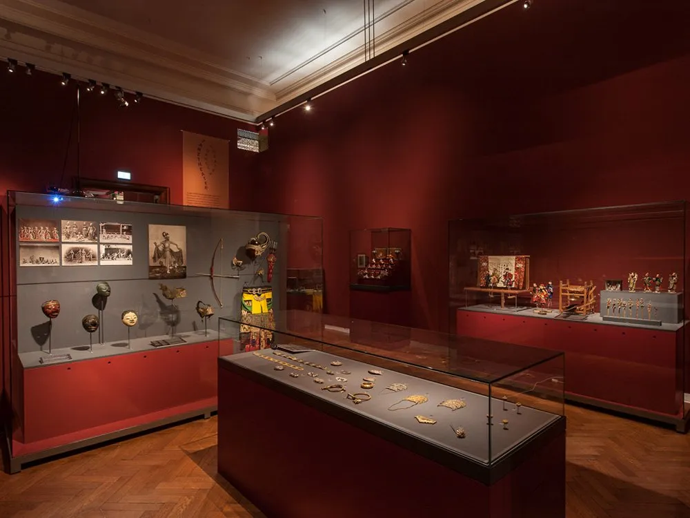 Ausstellungsraum: Vitrinen mit Masken, Schmuck, Puppen und historischen Fotos, beleuchtet mit gezielten Scheinwerfern