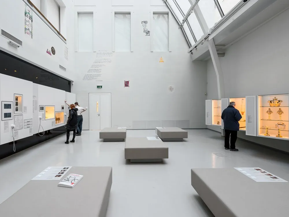 Ausstellungsraum mit grauen Sitzbänken, weißen Wänden mit Vitrinen, darunter eine mit goldenen Artefakten, und Besuchern, die die Ausstellungsstücke betrachten, unter einem Glasdach