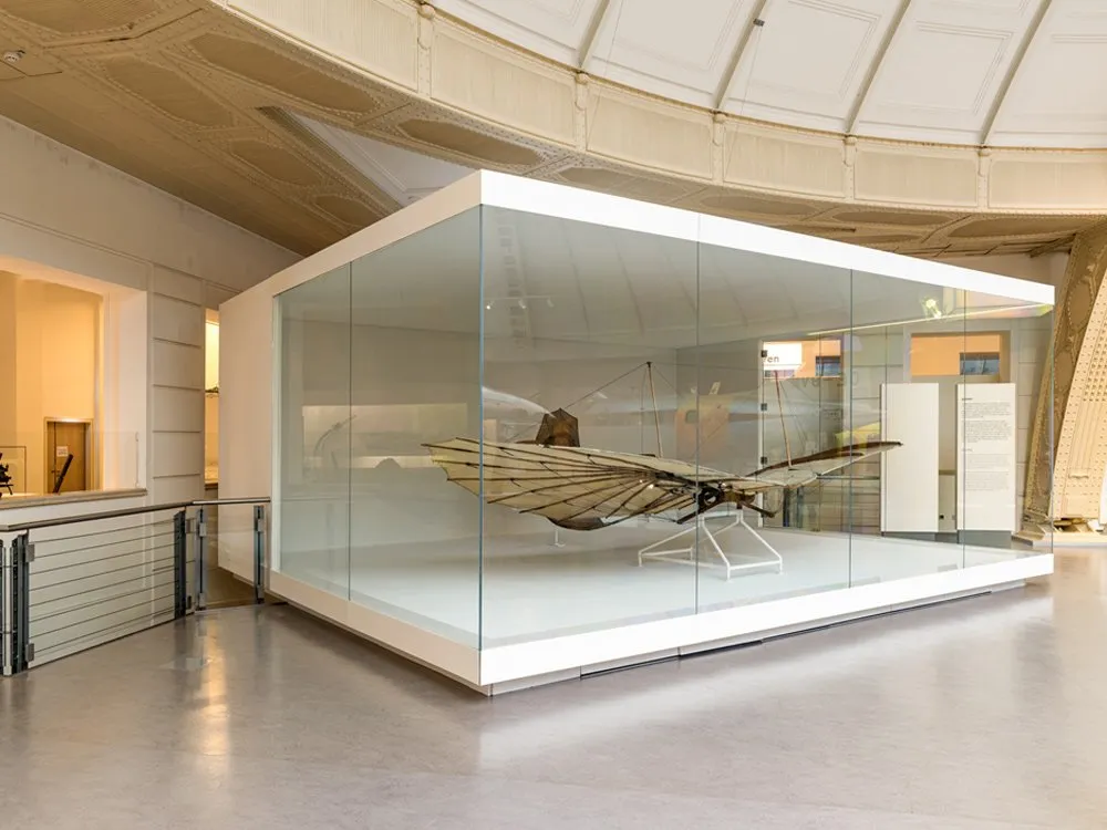 Ausstellungsraum: Helle Kuppelhalle mit einer gläsernen Vitrine, die ein Flugmodell mit Flügeln enthält, umgeben von Informationspaneelen.