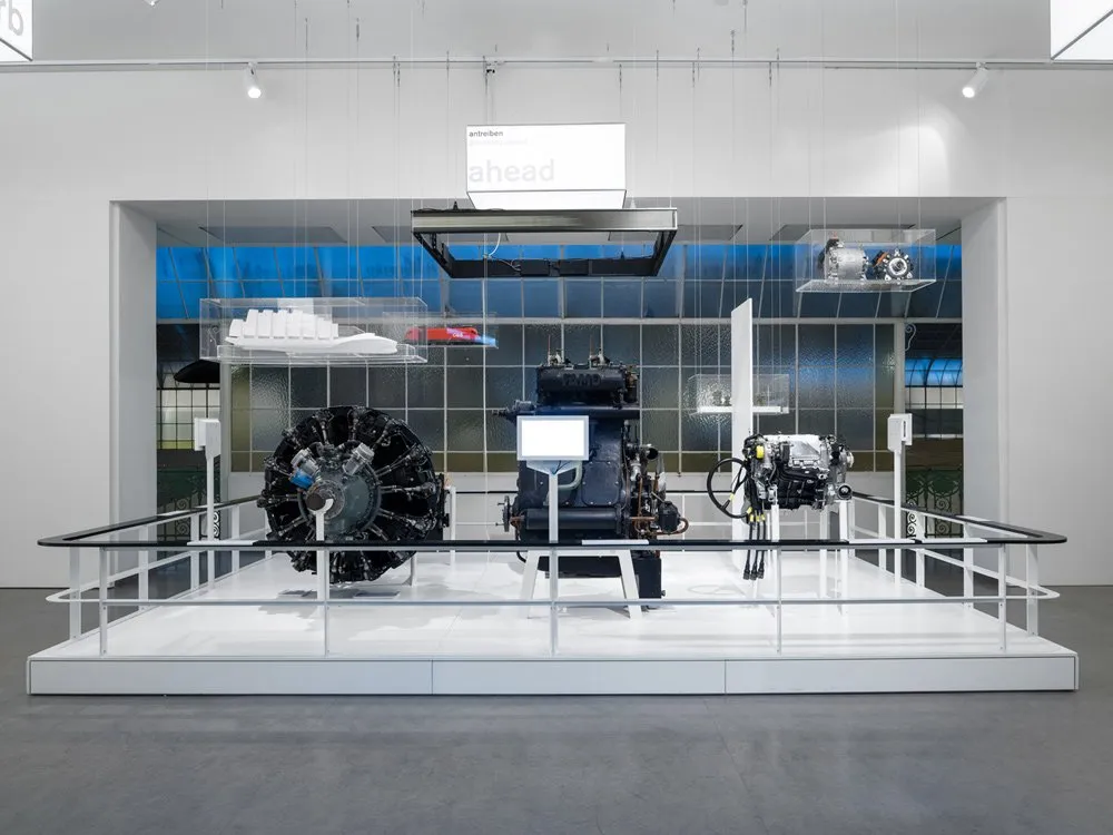 Ausstellungsraum: Weißer Raum mit einer Ausstellung von Motoren unter der Überschrift "an treiben ahead", umgeben von blauen Fenstern und Informationspaneelen.