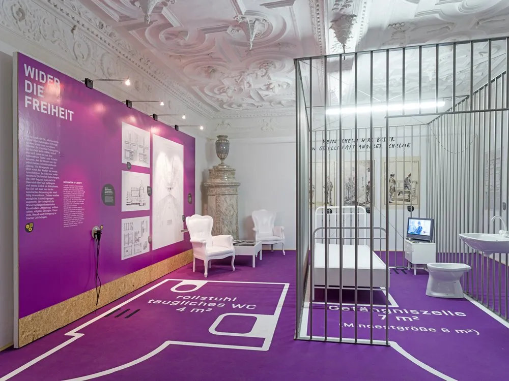 Ausstellungsraum: Lila Raum mit der Aufschrift "WIDER DIE FREIHEIT", einer Nachbildung einer Gefängniszelle mit Bett und Toilette sowie Informationspaneelen