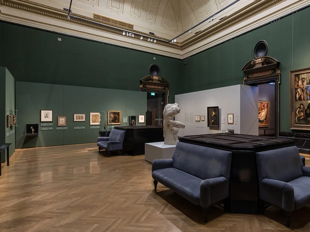 eleganter Saal mit grünen Wänden und hoher Decke. Gemälde und eine Skulptur einer Büste sind ausgestellt, umgeben von blauen Sesseln