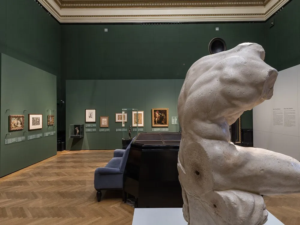 Ausstellungsraum: Ein Saal mit grünen Wänden und Holzfußboden. Eine große Skulptur einer Torso-Figur steht im Vordergrund, umgeben von Gemälden und blauen Sesseln.