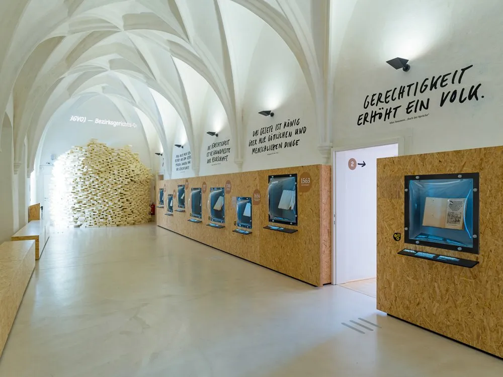 Ausstellungsraum: Gewölbter Raum mit der Aufschrift "GERECHTIGKEIT ERHÄLT EIN VOLK" und einer Holzstruktur mit Vitrinen, neben einem Stapel Büchern.