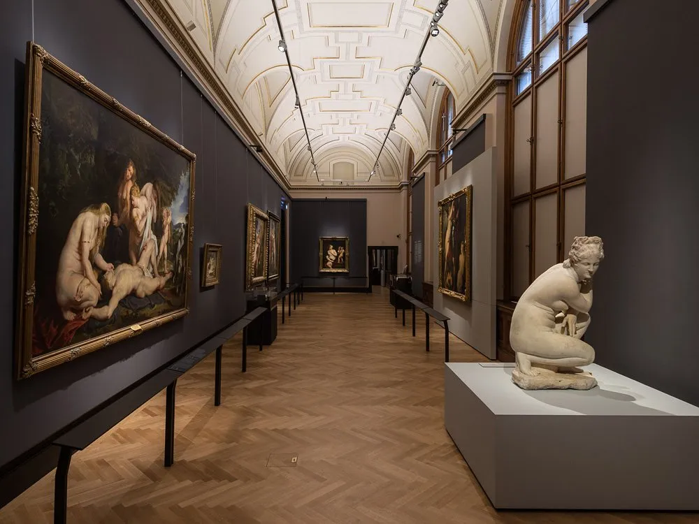 langer Saal mit hoher Decke und Holzfußboden. An den Wänden hängen Gemälde, eine Skulptur einer sitzenden Figur steht im Vordergrund, weitere Kunstwerke sind entlang der Wände ausgestellt.