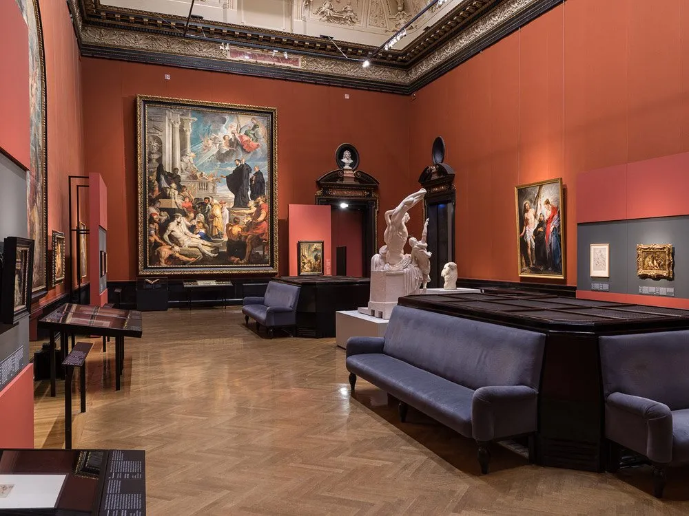 Saal mit orangen Wänden und hoher Decke. Große Gemälde hängen an den Wänden, eine Skulptur von kämpfenden Figuren steht im Zentrum, umgeben von blauen Sesseln