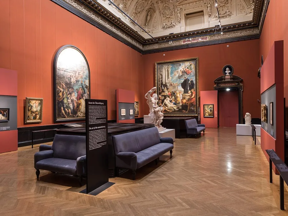 Saal mit orangen Wänden und hoher, verzierten Decke. Große Gemälde hängen an den Wänden, eine Skulptur von kämpfenden Figuren steht im Zentrum