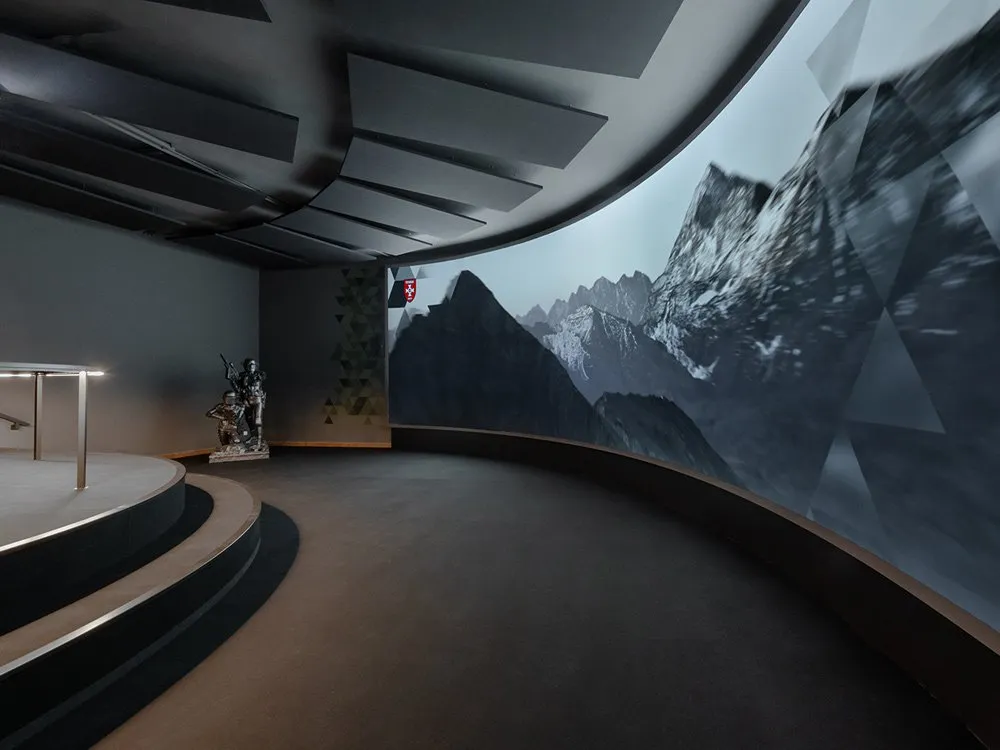 Ausstellungsraum: Dunkler Raum mit einer gebogenen Wand, die eine Berglandschaft mit Schweizer Flagge zeigt