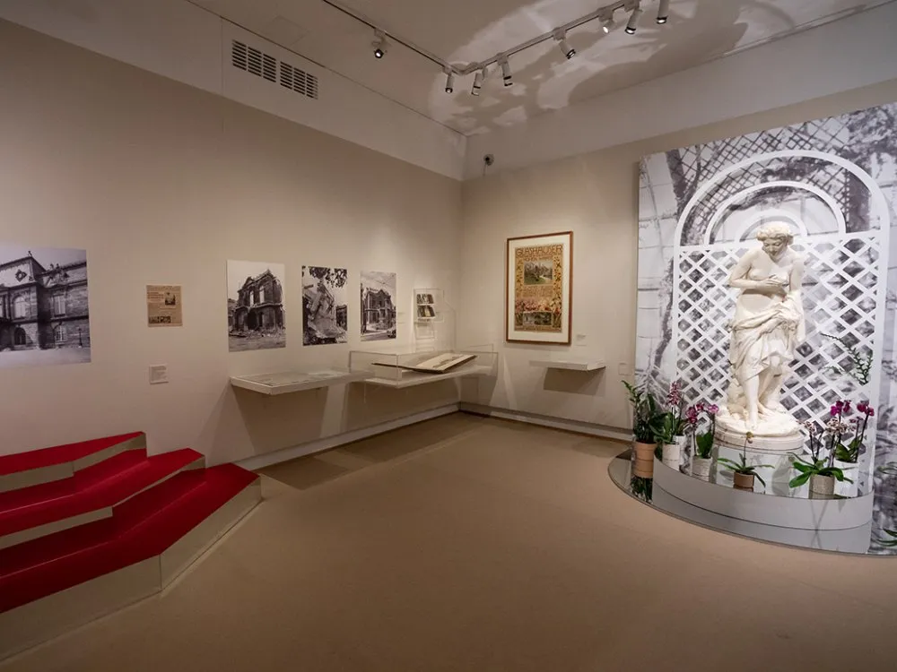 Ausstellungsraum: Weiße Statue einer Frau vor Gitterwand mit Pflanzen, alte Fotos, Poster und Vitrine