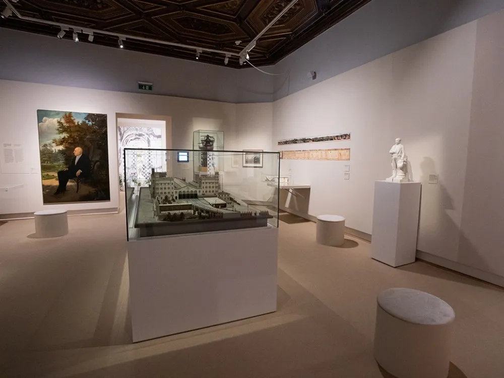 Ausstellungsraum: Porträt eines sitzenden Mannes, einer Modelllandschaft in einer Vitrine, einer weißen Statue und einem langen historischen Band an der Wand, umgeben von Sitzgelegenheiten.