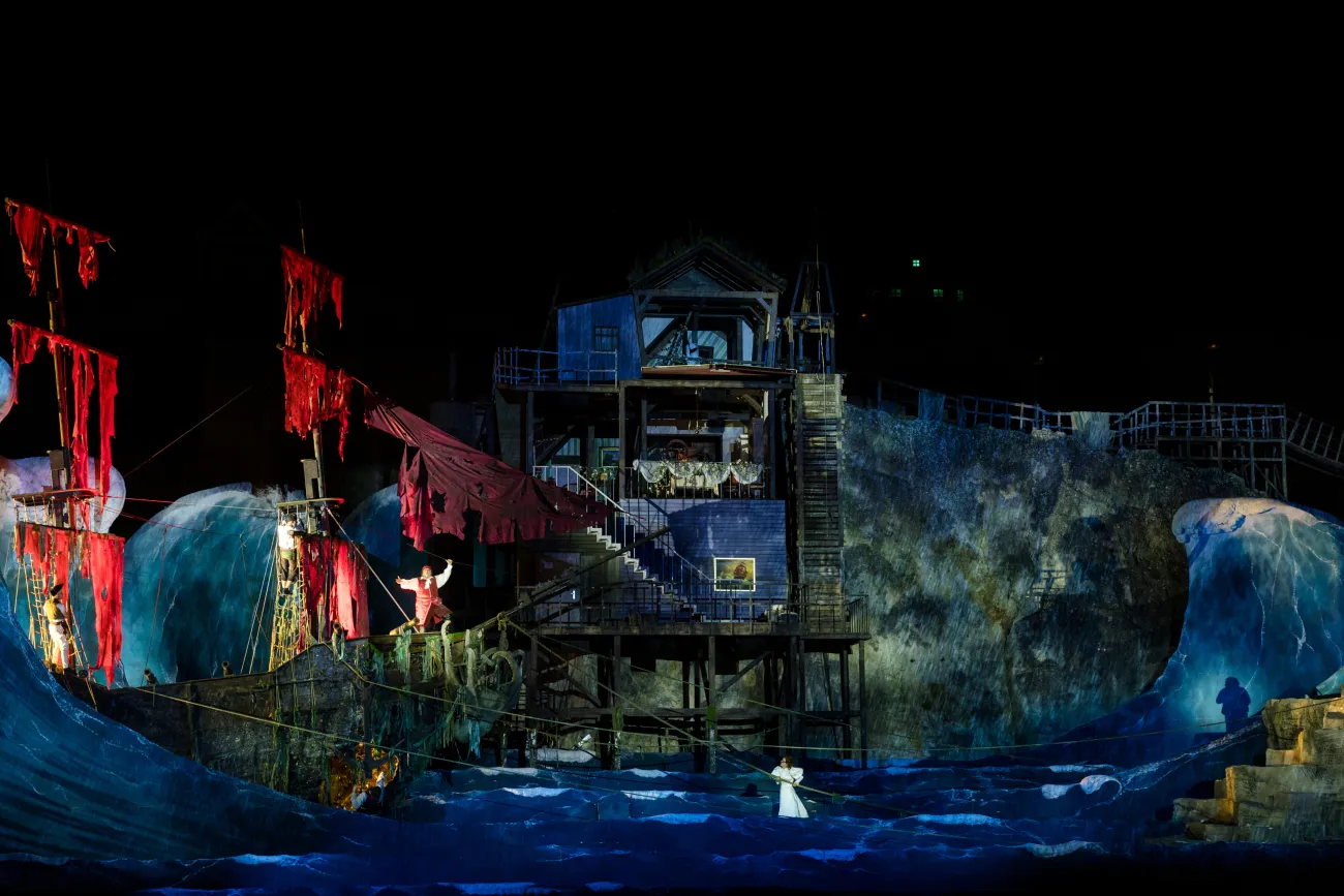 Szene: verwüstetes Schiff mit roten zerfetzten Segeln und stürmischen Wellen, beleuchtet von blauem Licht, Schauspieler auf dem Deck und im Wasser