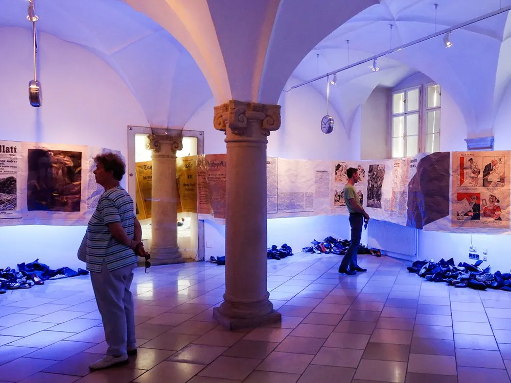 Gewölbter Raum mit Säulen, beleuchtet in Blau. Zwei Personen betrachten Ausstellungsplakate an den Wänden. Am Boden liegen Schuhe und Kleidungsstücke